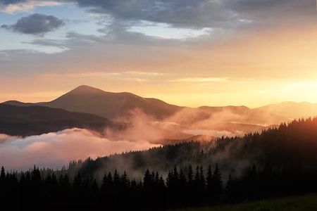 Sun setting behind a mountain range, its light breaking through orange clouds and fog in the valley
